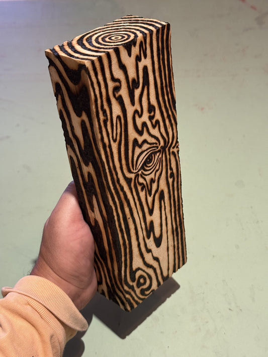 The tall block held in hand, an eye burned into the grain mid-face with concentric rings on the top end